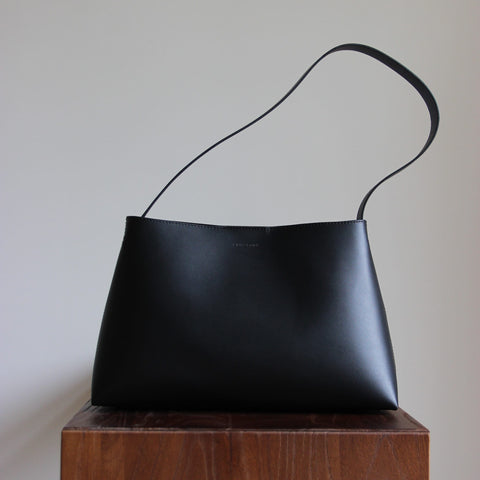 Schwarze Lederschultertasche aus pflanzlich gegerbtem Leder in Frontalansicht auf einem dunkelbraunen Echtholz-Kubus.