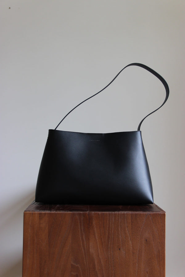 Schwarze Lederschultertasche aus pflanzlich gegerbtem Leder in Frontalansicht auf einem dunkelbraunen Echtholz-Kubus.