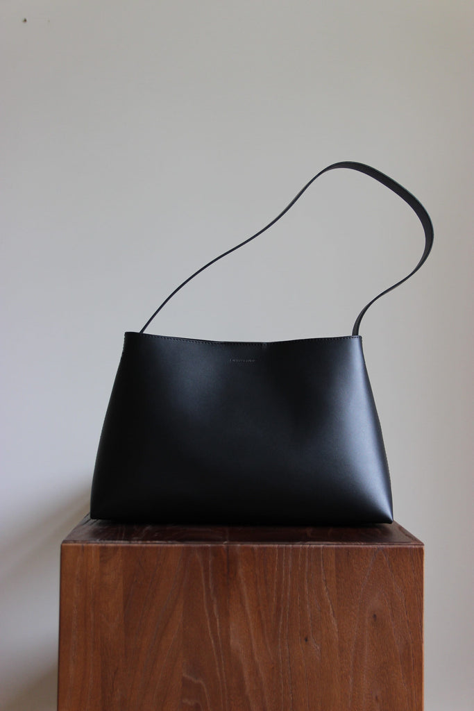 Schwarze Lederschultertasche aus pflanzlich gegerbtem Leder in Frontalansicht auf einem dunkelbraunen Echtholz-Kubus.